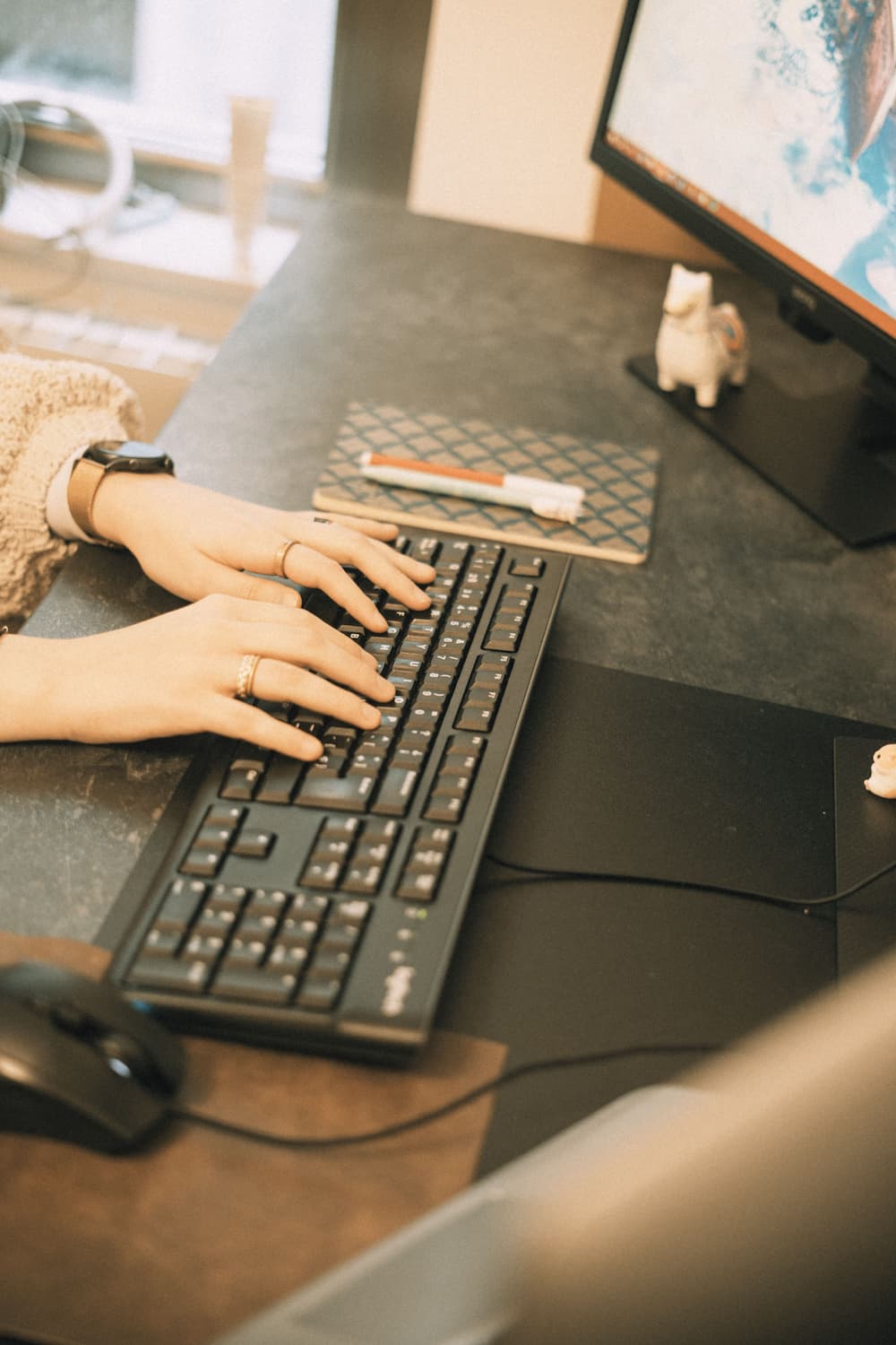 A woman’s hands are writing on a keyboard next to a mouse, a planner and two pens on a gray desk