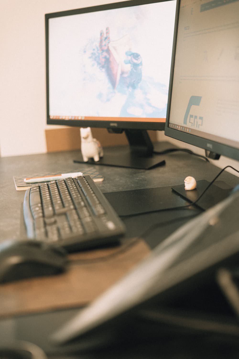 Two computer screens next to a keyboard, a mouse, a notebook and a toy llama on a gray desk