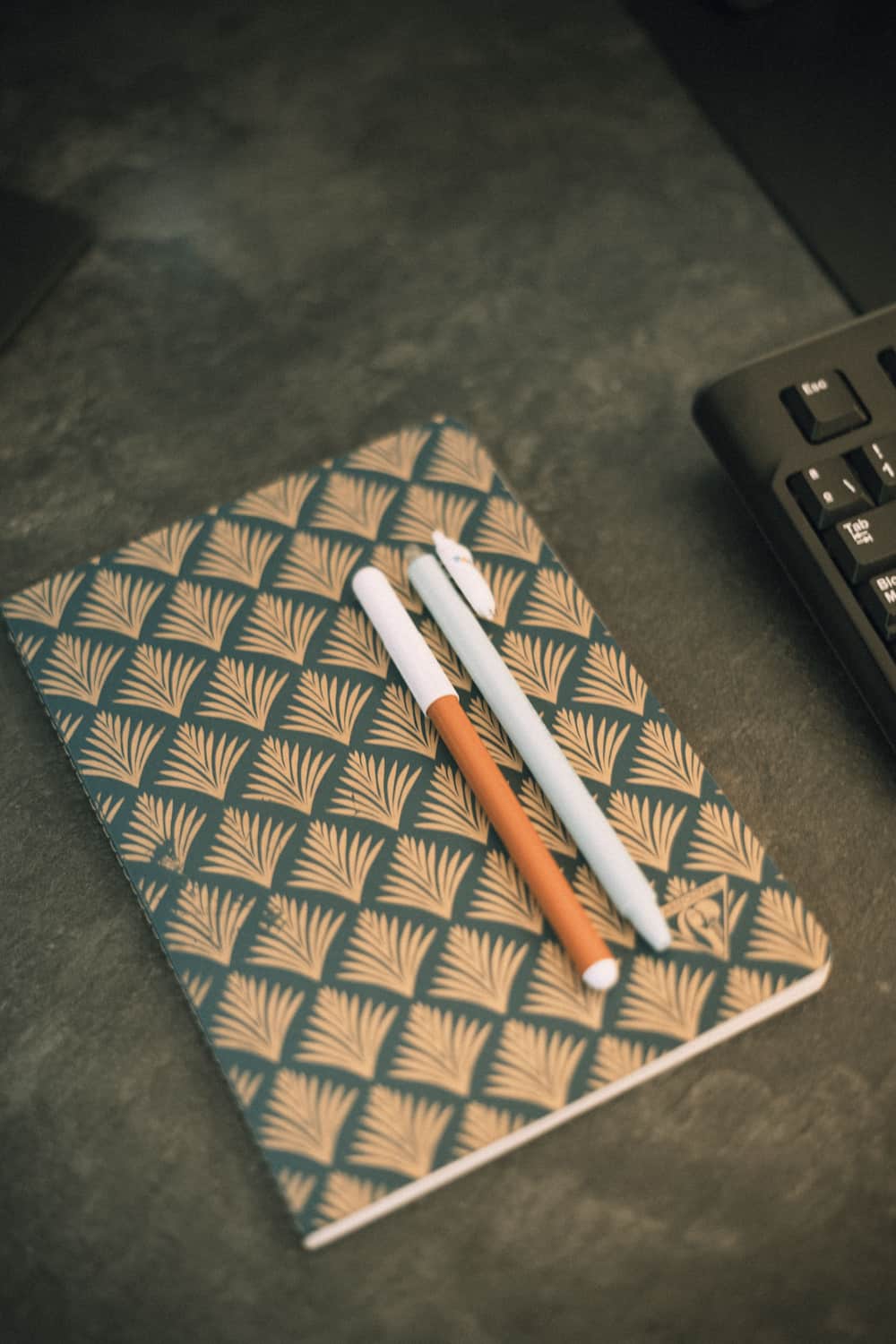 Close-up shot of a closed notebook with two pens on top of it next to a keyboard