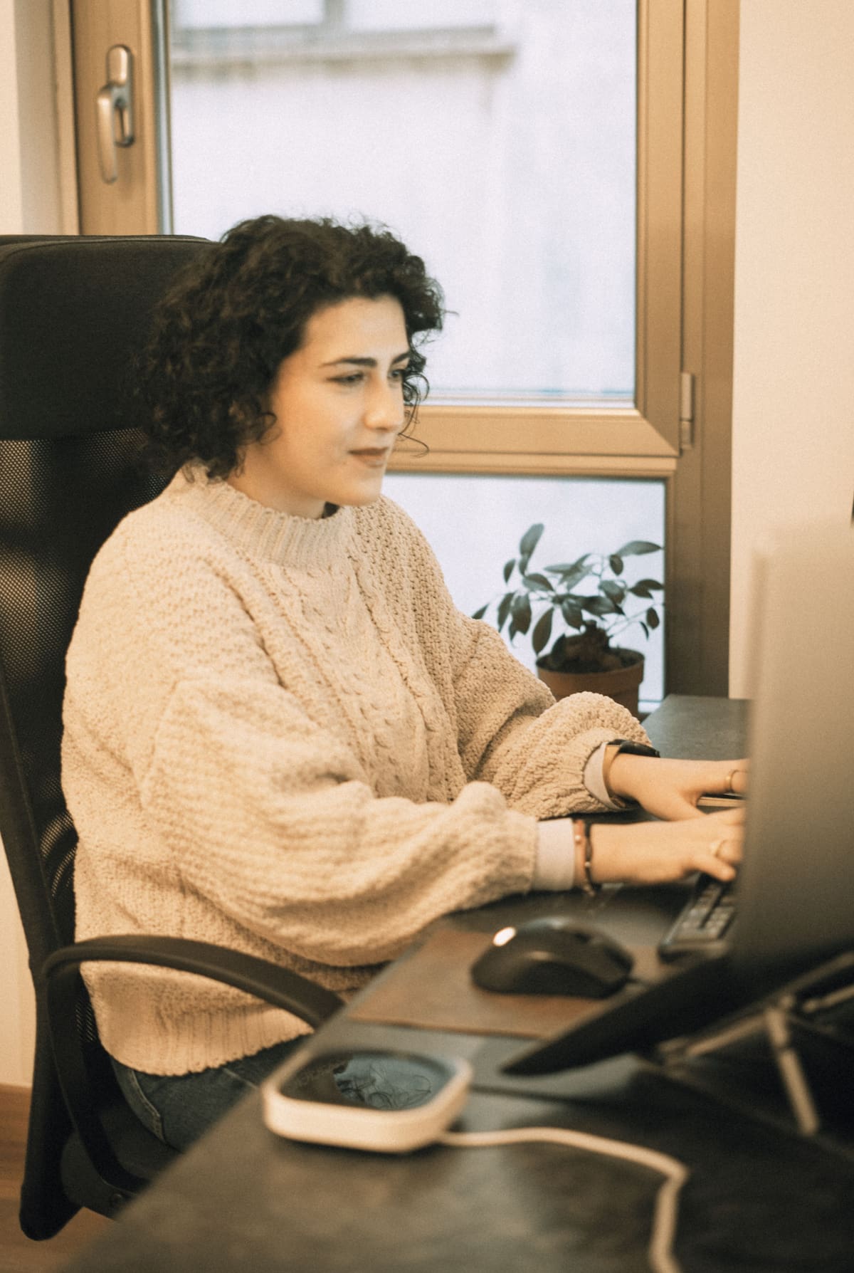 A woman is sitting while observing the secondary screen of a laptop and typing on a keyboard