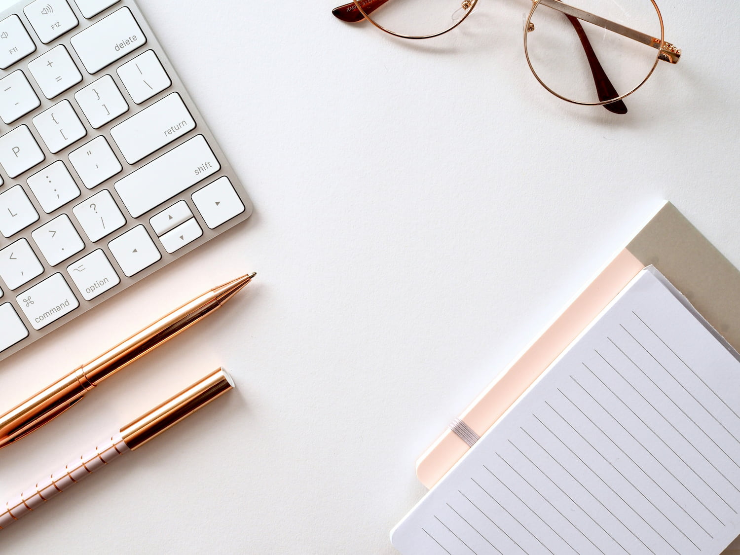 Close-up image of a white desk with a keyboard, a planner, two pens and a pair of glasses