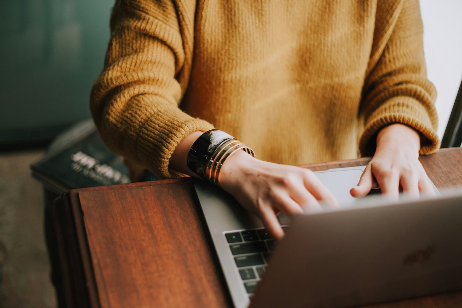 A woman with a mustard-colored sweater is writing on a keyboard on a wooden desk