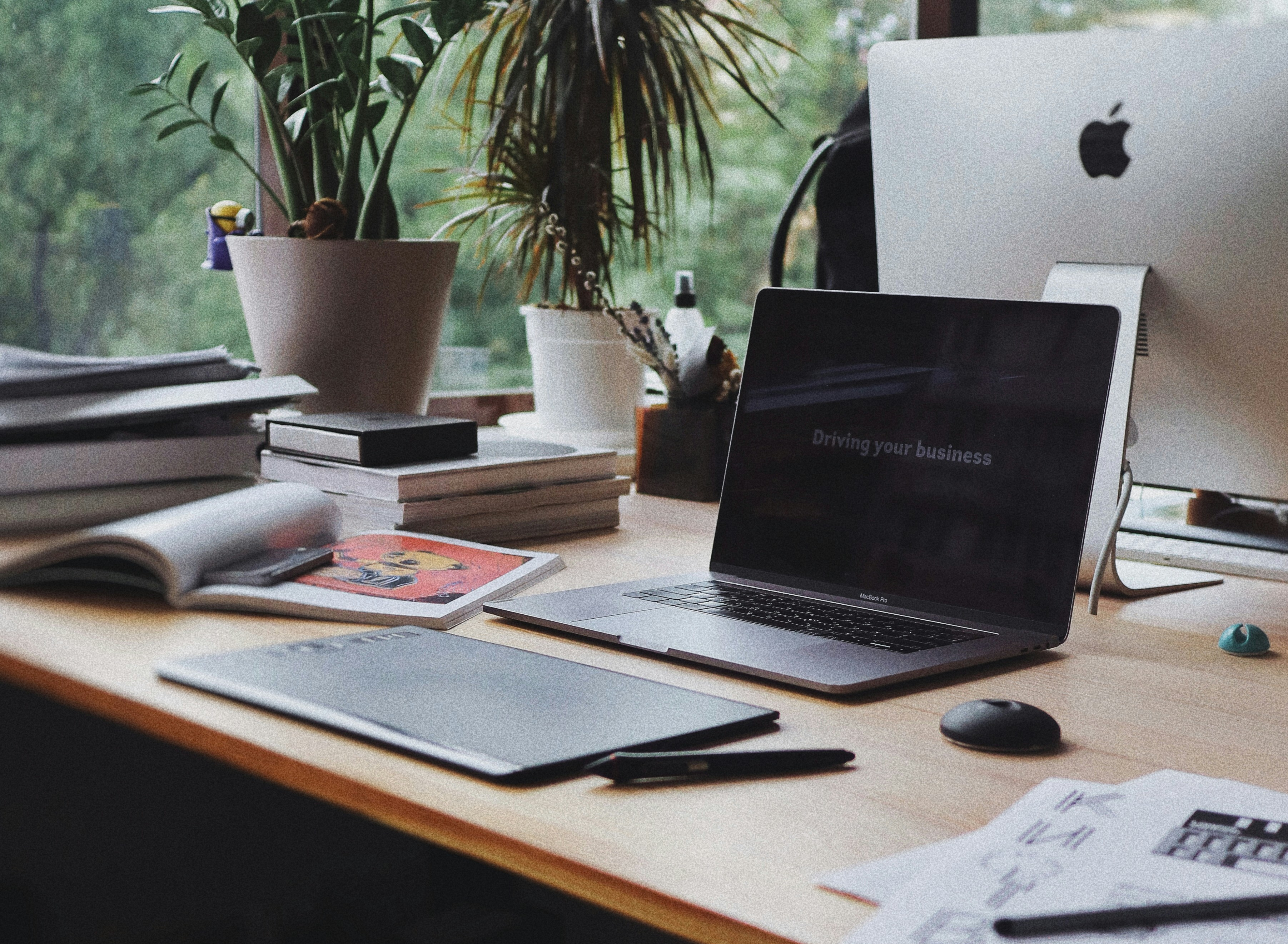 Close-up shot of a work desk with a computer, books, planners, pens and plants